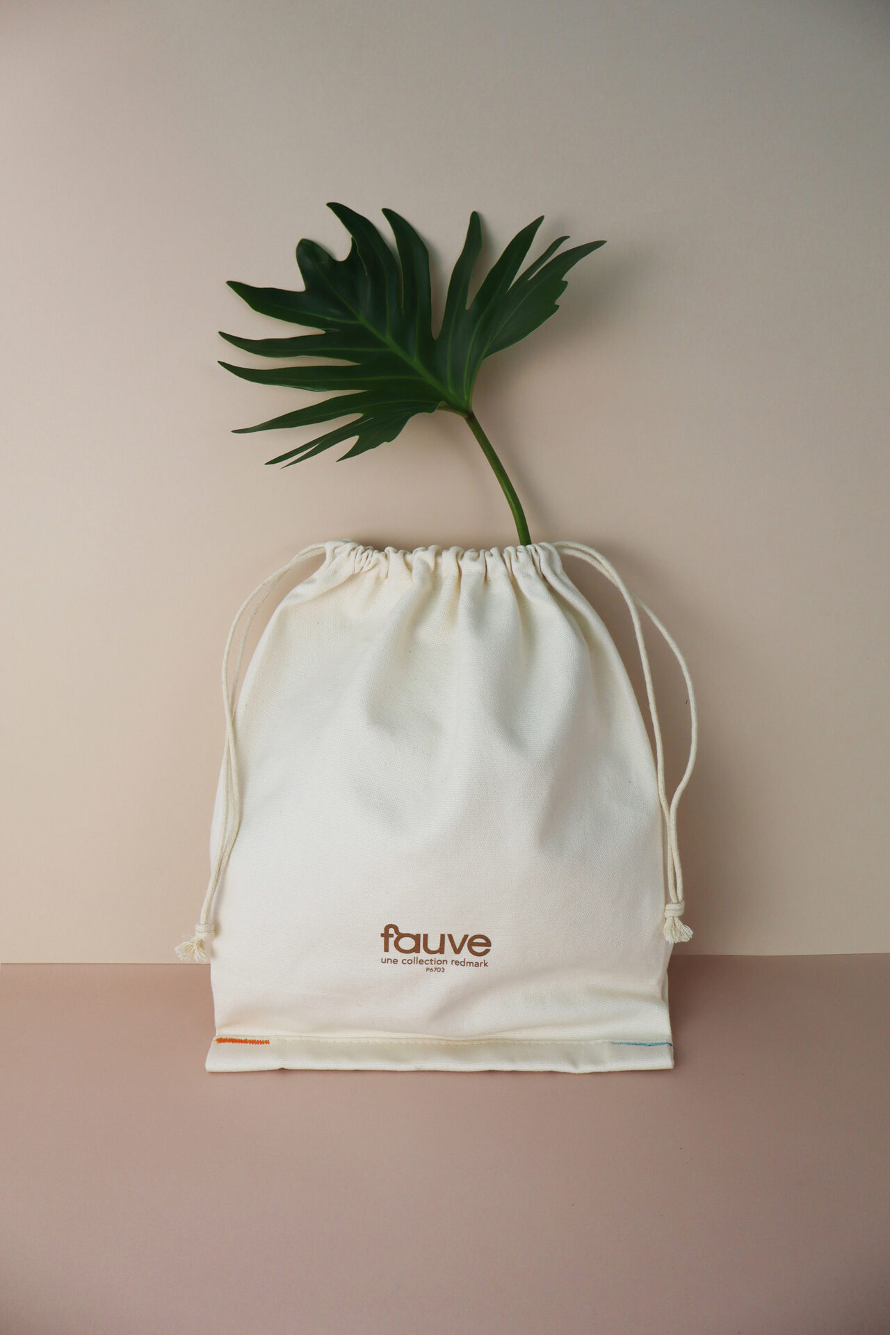 A white fabric bag with the 'fauve' logo on the front, with a tropical green leaf poking out from the top of the bag, against a simple, minimalist background of neutral tones.